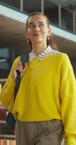Vertical Screen: Beautiful Female Student Walking in an Empty Public Library with Bookshelves. Young Smart Woman Carrying a Backpack with Textbooks to Prepare and Study for Exams. Low Angle Portrait