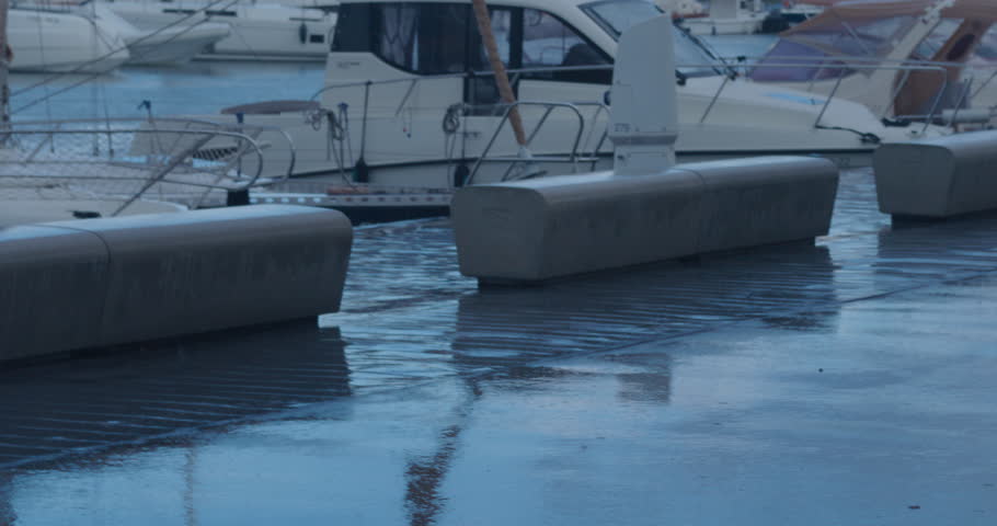 Handheld shot with view of the port in rain. Reflection on wet asphalt. Boats and yachts parked near pier. Summer or autumn rainy day. Moody atmosphere. Space for text. Overcast weather. 