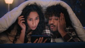 Gadget addict multiracial couple diverse man and woman scrolling mobile phone at night under blanket confused talking disagreement puzzled with smartphone error mistake shock bad news insomnia problem - Powered by Shutterstock - Get 15% off with code: PIKWIZARD15