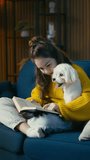 Teenage girl reading a book on sofa and petting lovely Maltese dog, friendship
