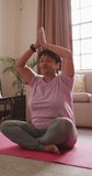 Vertical video of senior biracial woman practicing yoga and meditating in living room. healthy and active retirement at home.