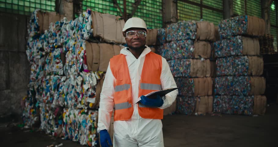 Portrait of a happy man with Black skin in a white uniform and an orange vest who stands near a huge pile of recycled plastic at a waste recycling plant and holds a tablet in his hands