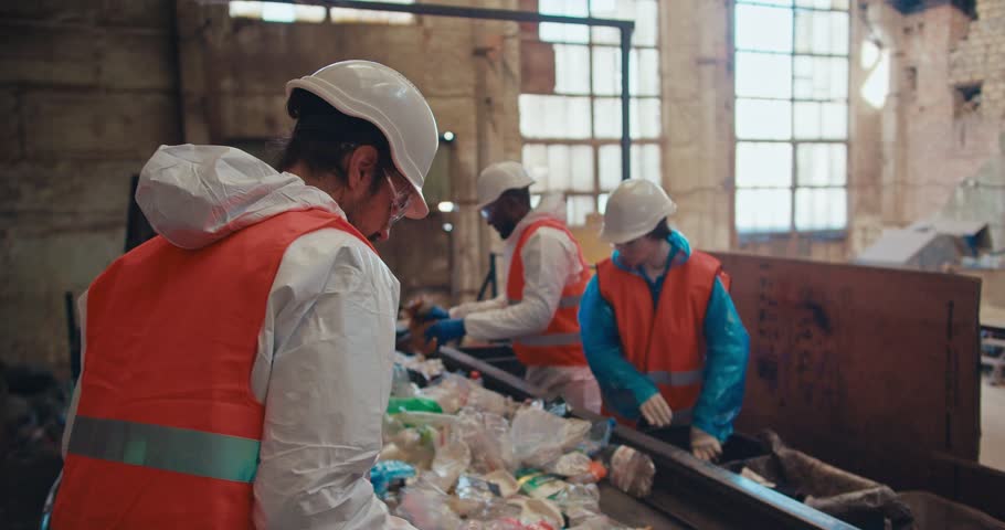 A brunette man in a special uniform and an orange vest recycles garbage and selects plastic bottles of a specific color along with his employees near a conveyor belt at a waste recycling plant