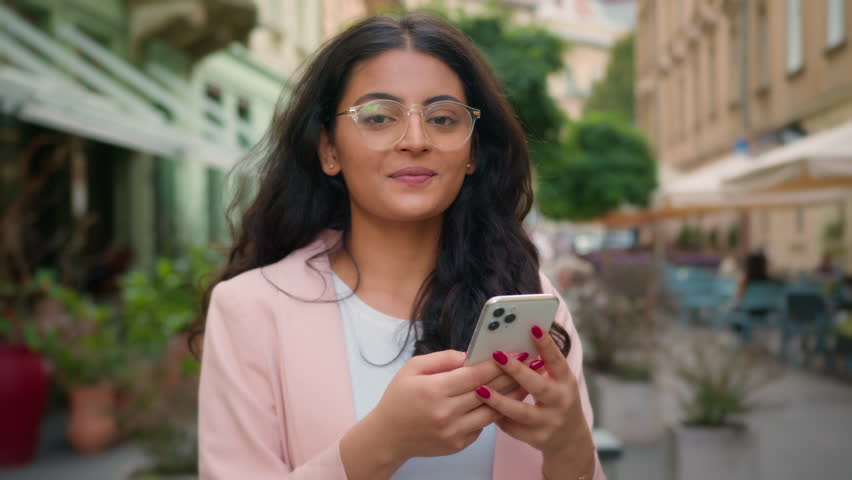 Happy successful smiling woman Indian Arabian ethnic female girl customer user holding mobile phone outside city street cafe. Positive client nodding head yes good reaction approve technology gadget Royalty-Free Stock Footage #3454940137