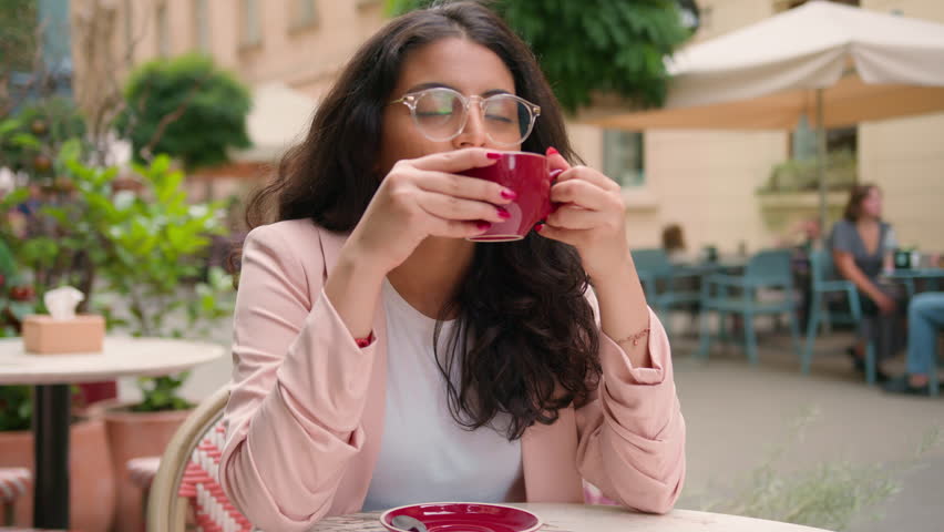 Morning relaxed businesswoman breakfast elegant woman Indian Arabian ethnic female girl client customer tasting drink sipping drinking coffee looking away weekend resting outside city cafe cafeteria