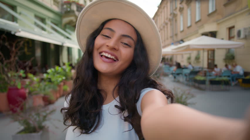 Young happy smiling tourist Indian Arabian lady woman female student waving hand social media vlogger holding smartphone talking camera shooting making online video call virtual mobile outside street