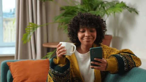 Good morning. African girl drinking coffee holding smartphone sitting on couch at home Woman with cell phone surfing internet using social media apps. Shopping online Internet news cellphone addiction Arkivvideo