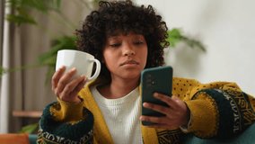 Good morning. African girl drinking coffee holding smartphone sitting on couch at home Woman with cell phone surfing internet using social media apps. Shopping online Internet news cellphone addiction - Powered by Shutterstock - Get 15% off with code: PIKWIZARD15