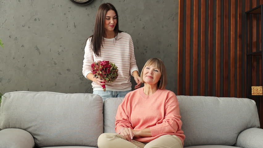 Two adult women older mature mom young kid receiving flowers hugs kiss sits on gray sofa couch stay home flat rest relax spend free spare time in living room indoor. Mother's Day love family concept Royalty-Free Stock Footage #3454981247