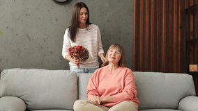 Two adult women older mature mom young kid receiving flowers hugs kiss sits on gray sofa couch stay home flat rest relax spend free spare time in living room indoor. Mother's Day love family concept - Powered by Shutterstock - Get 15% off with code: PIKWIZARD15