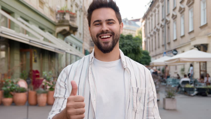 Young Caucasian man posing smiling showing thumb up happy joyful cheerful guy enjoying toothy dental smile satisfaction funny urban city outside businessman friendly male model gesture positive sign