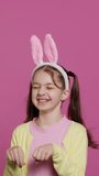 Vertical Video Joyful carefree schoolgirl jumping around in studio, imitating a rabbit and hopping against pink background. Cheerful active child wearing bunny ears and bouncing, adorable kid. Camera