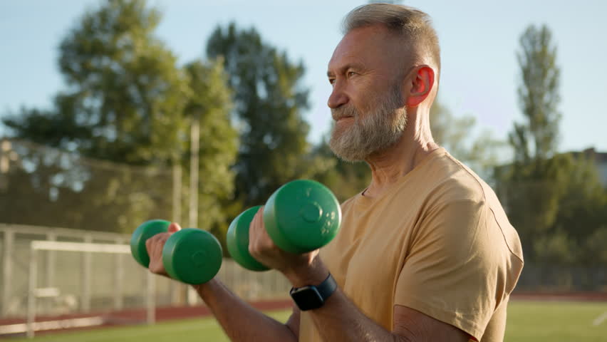 Elderly Caucasian pensioner man lift dumbbells confident sportsman athlete training physical activity workout stadium city outside muscles bodybuilder strong exercise powerful arms biceps heavy weight