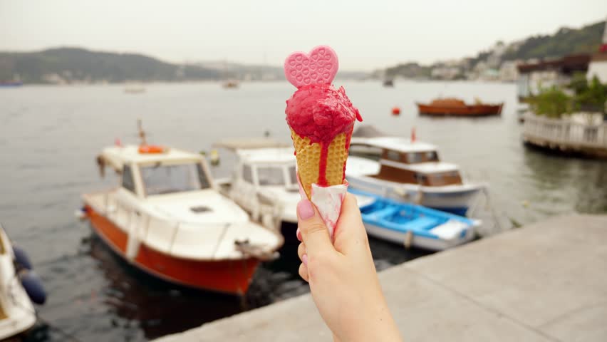 Tourist woman holds pink ice cream cone with heart-shaped cookie, standing at waterfront in Bebek, Istanbul. First-person view with close-up focus on melting gelato, blurred fishing boats rock on back
