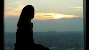 Woman sits by window in dimly lit room, gazing out at city in late evening. Atmosphere is introspective, capturing the mood of uncertainty and solitude. Cityscape seen outside below in twilight - Powered by Shutterstock - Get 15% off with code: PIKWIZARD15