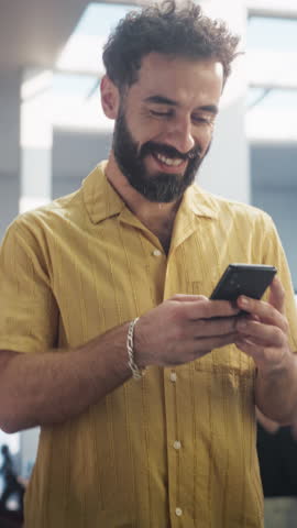 Vertical Screen: Portrait of Young Man in Casual Clothes Walks in Corporate Office Hallway, Using his Smartphone and Smiling. Successful Businessman Communicating with Colleagues Over Text Messages