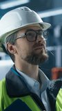 Vertical Screen: Portrait of Caucasian Male Production Supervisor Wearing Glasses and Looking at Camera, Smiling. Technician in Hardhat And Reflective Jacket at Factory with Robotic Arms Assembly Line