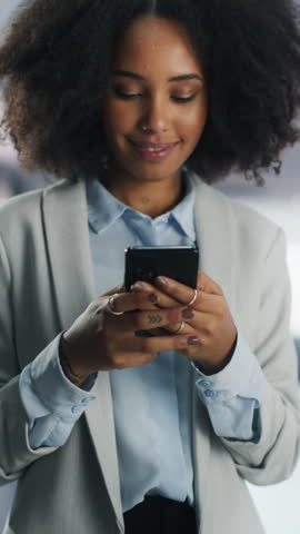 Vertical Screen: Smiling Young Black Woman Browsing Internet and Social Networks on Her Smartphone. Young Female Manager Checking Dating Apps, Shopping Online, Messaging Friends and Family Royalty-Free Stock Footage #3455193283
