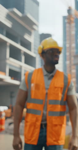 Vertical Screen: Portrait of an African Builder Wearing a Hardhat and Safety Vest while Posing for Camera and Smiling at a Commercial Building Construction Site. Black Man Crossing His Arms Royalty-Free Stock Footage #3455207521