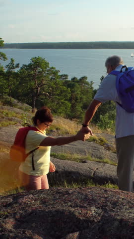 Elderly active couple climbing on the rock and admiring the sunrise, the sunset. Scandinavian landscape with sea and pine trees.