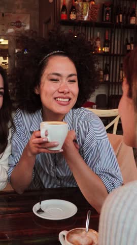 Vertical portrait of a young women talking and laughing sitting on a coffee shop or restaurant. Two ladies smiling and looking his friend holding a cup. Girls having fun chatting on a bar. High