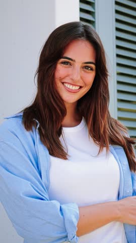 Vertical portrait of happy teenage brunette girl standing outdoors with adorable perfect white teeth smile. Slow motion. One young beautyful woman smiling and looking at camera with crossed arms. High