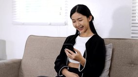 Young Asian woman happily playing with mobile phone and laughing in living room at home.