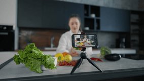 Young woman video lesson about cooking. Positive girl talks recording video lesson, filming cooks vegetables tutorial in kitchen. Concept for online healthy cooking courses, recording of training.