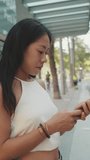 Vertical video, Cute brunette girl dressed in white top and jeans looks around at public transport stop, looks into smartphone