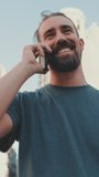 VERTICAL VIDEO, Smiling young man with beard talking on the phone on modern buildings background
