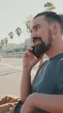 VERTICAL VIDEO, Young man with beard sits on bench talking on cellphone