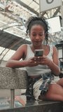 VERTICAL VIDEO, Young woman sitting at the railway station, using cellphone