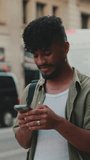 VERTICAL VIDEO: Young smiling man with beard dressed in an olive-colored shirt uses phone