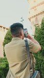 VERTICAL VIDEO: Close up, young man dressed in an olive-colored shirt takes photo on the street of the old city. Back view