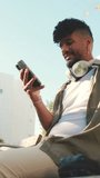 VERTICAL VIDEO: Young man with beard wearing an olive-colored shirt with headphones sits on bench and uses cellphone