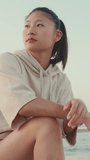 VERTICAL VIDEO, girl wears sportswear sits on the promenade at morning time, on the background of the sea