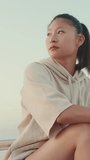 VERTICAL VIDEO, girl wears sportswear sits on the promenade at morning time, on the background of the sea,turns her head and looks at the camera