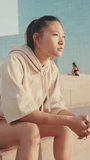 VERTICAL VIDEO, girl in sportswear resting after workout while sitting on steps on modern buildings background at sunrise. Young woman turns her head and looks at the camera
