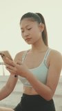 VERTICAL VIDEO, girl in sports top sits on the waterfront, uses smartphone