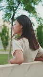 Vertical video: Beautiful brunette girl is resting sitting on bench in city park