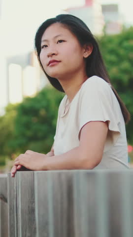 Vertical video: Young thoughtful woman stands leaning on railing and looks into the distance