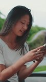 Vertical video: Beautiful happy brunette girl dressed in casual clothes, uses mobile phone while sitting on bench outside, checking news, texting messages