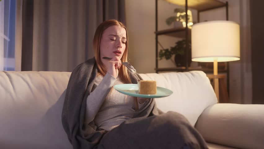 Portrait of sad young blond woman dealing with stress by eating food sitting on sofa late at home Upset female wrapped in blanket crying and eating cake at night indoors alone Mental heath problem Royalty-Free Stock Footage #3455663869