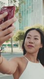 Vertical video, Cute happy girl dressed in white top and jeans makes selfie on mobile phone while standing on modern city background