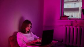 young woman lying in bed with laptop in neon light at home, working online at home training and online consultation.