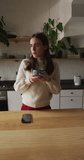 Vertical video of caucasian non-binary transgender woman drinking coffee in kitchen. spending quality time at home alone, body inclusivity.