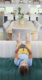 Vertical video of happy caucasian boy lying on sofa and using smartphone, slow motion. Spending quality time, domestic life and childhood concept.