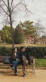 Vertical video, Happy young couple using smartphone together outdoors on bench, sharing moment of connectivity and leisure.