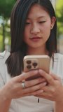 Vertical video: Close up, beautiful young woman sits on bench in city park using mobile phone, blurred foreground