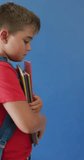 Vertical video of happy caucasian boy holding books on blue background, copy space, slow motion. Childhood, education, learning and background concept.